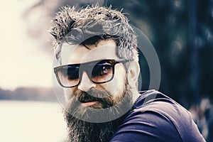 Man with beard and mustache wears sunglasses, river embankment on background. Hipster on confident face with fashionable