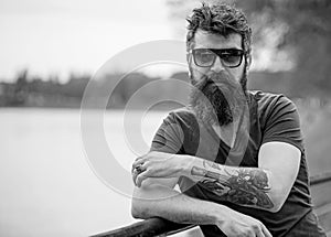 Man with beard and mustache wears sunglasses, river embankment on background. Hipster on confident face with fashionable