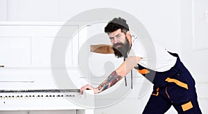 Man with beard and mustache, worker in overalls pushes piano, white background. Courier delivers furniture in case of