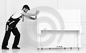 Man with beard and mustache, worker in overalls pushes piano, white background. Courier delivers furniture in case of