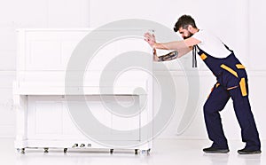 Man with beard and mustache, worker in overalls pushes piano, white background. Courier delivers furniture in case of