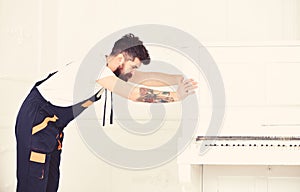 Man with beard and mustache, worker in overalls pushes piano, white background. Delivery service concept. Loader moves