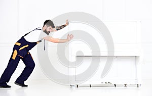 Man with beard and mustache, worker in overalls pushes piano, white background. Loader moves piano instrument. Courier