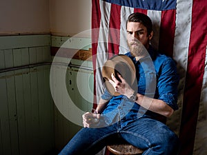 Man with beard sitting against an American flag in a dirty room