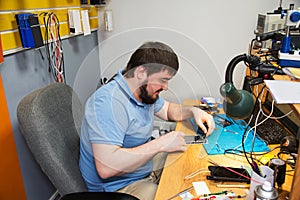 A man with a beard sitting at a table looks at a disassembled cell phone, repair of equipment telephony modern
