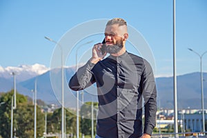 Man with a beard and slicked back hair in black shirt talking on phone, stands on the street of the city from behind against the