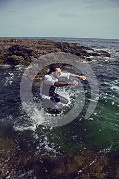 man with a beard and sunglasses in clothes a vest and a white shirt jump in sea among the rocks in summer