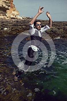 man with a beard and sunglasses in clothes a vest and a white shirt jump in sea among the rocks in summer