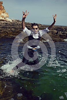 man with a beard and sunglasses in clothes a vest and a white shirt jump in sea among the rocks in summer