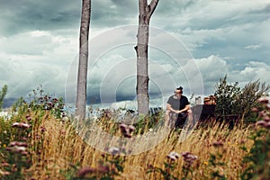 Man with beard and sunglasses sitting on the mountain top on wooden bench, surrounded with tall grass and wild flowers