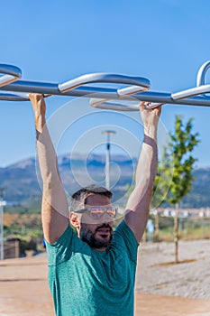 man with beard and sunglasses training his back by doing pull-ups on a bar in an outdoor gym