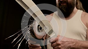 Man with beard and tattoo in T-shirt in black studio, sharpen a machete. Closeup