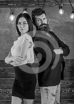 Man with beard and teacher in eyeglasses stand back to back, chalkboard on background. School staff concept. Lady and