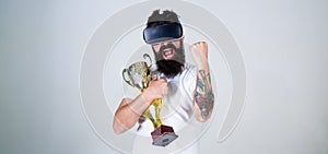 Man with beard in virtual reality glasses holds goblet, light background. Hipster on happy face squeezing fist as