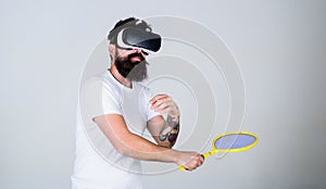 Man with beard in VR glasses beating pitch, grey background. Guy with VR glasses play tennis with racket. Virtual sport