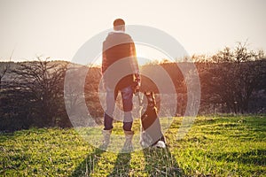 A man with a beard walking his dog in the nature, standing with a backlight at the rising sun, casting a warm glow and