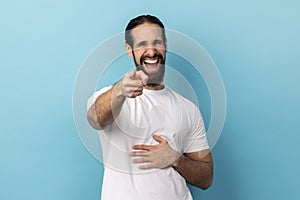 Man with beard wearing white T-shirt pointing index finger at camera and laughing positively.