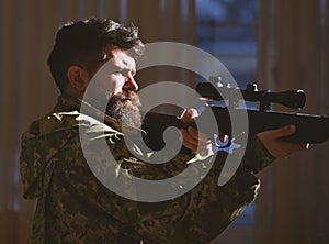 Man with beard wears camouflage clothing, dark interior background. Macho on suffering grimace face aiming at victim