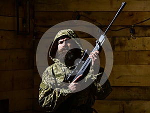 Man with beard wears camouflage hooded clothing, wooden interior background. Macho on strict face at gamekeepers house