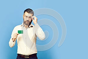 Man with beard in white shirt holds green business card. Guy with smart face with glasses isolated on blue background