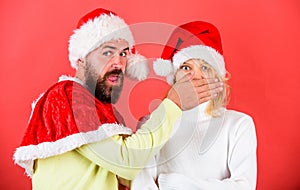 Man with beard and woman in santa hat on red background. Man covers mouth of girl to keep secret. Couple celebrate