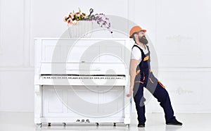 Man with beard worker in helmet and overalls pushes, efforts to move piano, white background. Courier delivers furniture