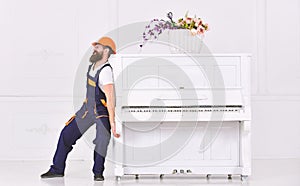 Man with beard worker in helmet and overalls pushes, efforts to move piano, white background. Courier delivers furniture