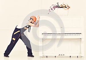 Man with beard worker in helmet and overalls pushes, efforts to move piano, white background. Heavy loads concept