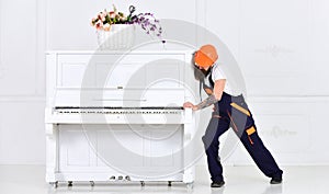 Man with beard worker in helmet and overalls pushes, efforts to move piano, white background. Loader moves piano
