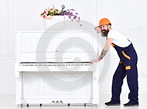 Man with beard worker in helmet and overalls pushes, efforts to move piano, white background. Loader moves piano