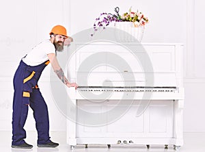 Man with beard worker in helmet and overalls pushes, efforts to move piano, white background. Loader moves piano