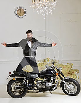 Man, bearded biker in leather jacket near motor bike in luxury living room interior background. Hipster biker brutal in