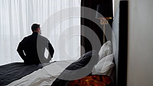 A man on a bed looks out the window, rear view. Young man waking up in the morning. In the morning, the guy sits with