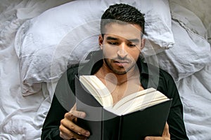 Man in bed with open shirt and pecs reading hardback book