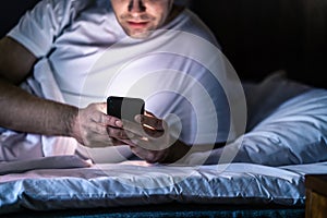 Man in bed with phone at night. Texting with smartphone before sleeping. Guy holding cellphone in hand. Dark home bedroom.