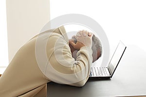 A man bends his head over a computer in despair while he puts his hands over his head. - Image