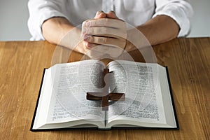 Man with Bible and cross praying at wooden table, closeup