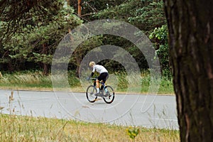 A man in a bicycle ride rides on an asphalt road in the woods and looks at the camera