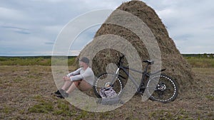 A Man With A Bicycle Sits Near A Haystack, Taking A Rest. Healthy Man with Cycling. Free Traveler Travels by Bike