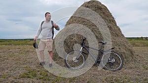 A man on a bicycle stops near a haystack in a field to take a break and relax. He Climbs Off The Bicycle, Leans It On A