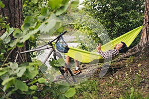 Man on bicycle trip at camping by lake is relaxing in green hammock while listening to music. Active recreation theme in