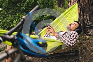 Man on bicycle trip at camping by lake is relaxing in green hammock while listening to music. Active recreation theme in