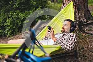 Man on bicycle trip at camping by lake is relaxing in green hammock while listening to music. Active recreation theme in