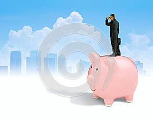 Man with binoculars standing on piggy bank seeking financial success with city background photo