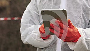 Man in bio-hazard suit and gas mask takes notes in his tablet standing on the polluted land