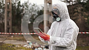 Man in bio-hazard suit and gas mask takes notes in his tablet standing on the polluted land