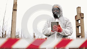 Man in bio-hazard suit and gas mask takes notes in his tablet standing on the polluted land