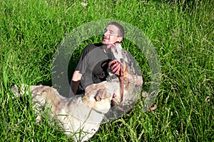 Man in black clothes with two dogs sitting on green grass, one dog licking his cheek