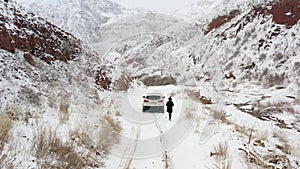 A man in a black coat with a backpack approaches a white car. Beautiful snow-covered mountain road