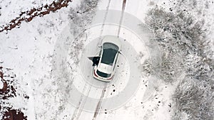 The man in black gets into a white car and starts driving. A view from a height of an SUV on a snowy mountain road
